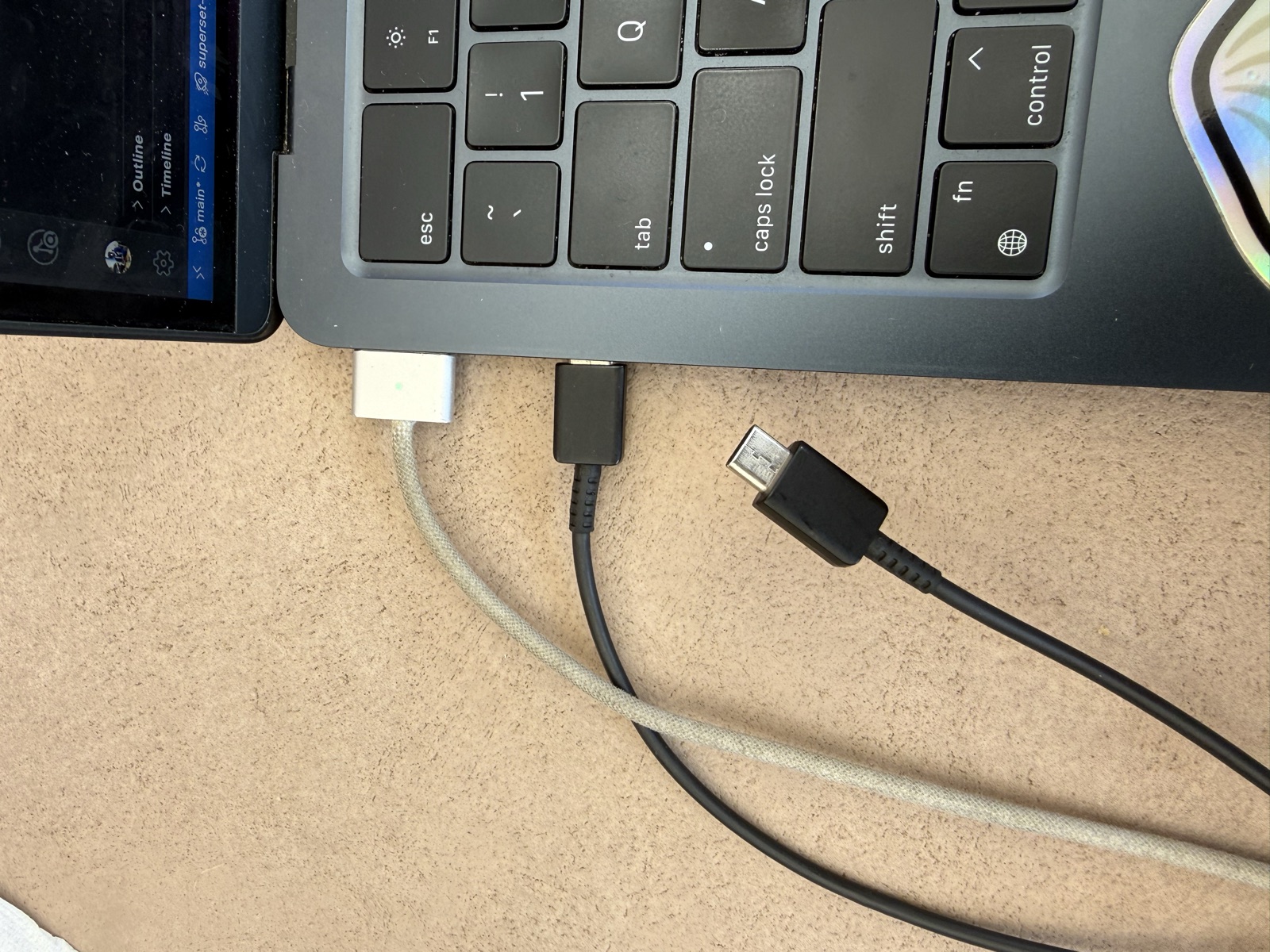 A MacBook on a cafe table with a USB-C cable next to its two left-side ports, not yet connected. The MagSafe charger is held away from the laptop.